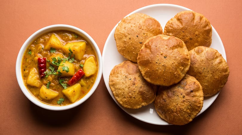 A bowl of potato curry with red chilies and cilantro, next to a plate of fried puri bread.