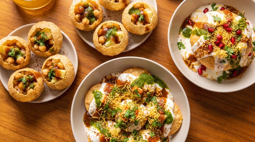 Three plates of Indian chaat dishes, including pani puri, dahi puri, and raj kachori, on a wooden table.