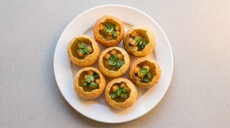 Seven pani puri, filled with chickpeas, green liquid, and cilantro, arranged on a white plate.