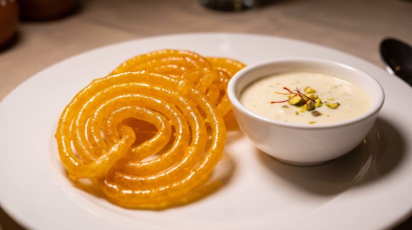 Golden jalebi spirals on a white plate next to a bowl of creamy rabri topped with pistachios and saffron.